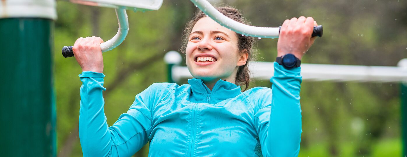 Smiling teenage girl exercising in the park, holding onto outdoor gym equipment wet with rain and lifting her body up, wearing turquoise waterproof